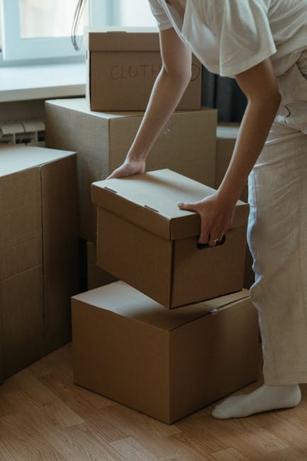 A person wearing light clothing and white socks is engaged in packing or unpacking cardboard boxes during a home relocation within a room that has abundant natural light from a large window. The indiv