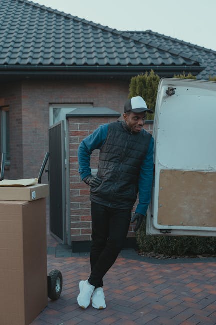 A person wearing light clothing and white socks is engaged in packing or unpacking cardboard boxes during a home relocation within a room that has abundant natural light from a large window. The indiv