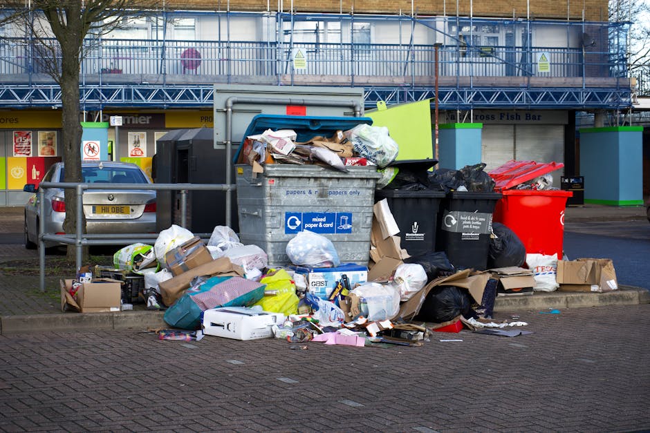 A large collection of mixed waste and recycling bins placed on a paved area in front of a building, with several bins overflowing with cardboard boxes, plastic bags, paper, and packaging materials. The scene includes a grey recycling container labeled for mixed paper and cardboard, a black waste bin, and a red recycling bin, all positioned close together. Some waste such as flattened cardboard, crumpled paper, and plastic packaging is scattered around the bins on the pavement. Behind the bins, there is a metal railing, with parked cars visible on the left side. In the background, a building with a scaffolding structure and a blue and green storefront is visible, along with a sign indicating rules for waste disposal at this location. The image captures a typical urban environment where rubbish is temporarily stored before collection, relevant to the logistics of house removals and furniture transport managed by companies like Removal Company Hounslow, especially when coordinating waste disposal during home relocations near Hounslow Central Station.