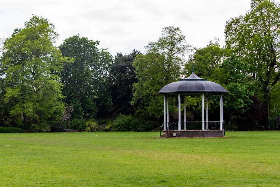 A gazebo with a dark, domed roof supported by white columns is situated on a brick foundation in the middle of a large, open grassy area within Lampton Park. The park features an expansive lawn with well-maintained, lush green grass extending into the distance. Surrounding the open space are numerous trees with vibrant green leaves, some with dense foliage and others less so, creating a rich backdrop of natural greenery. The sky overhead is partially overcast with light gray clouds, providing diffused lighting across the scene. In the context of house removals and furniture transport, this outdoor setting could be a staging or loading area, with potential for removal vans or trolleys nearby to facilitate the easy movement of packed boxes, furniture, or appliances during a home relocation. The environment appears calm and suitable for efficient packing and loading processes, consistent with professional moving services offered by Removal Company Hounslow in this area.