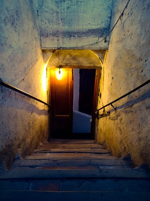 This image depicts a narrow, steep staircase inside a building, constructed from worn stone or concrete with visible cracks and uneven surfaces. Metal handrails run along both sides of the staircase, attached to the walls which are painted in a faded light color with signs of age and dirt. At the top of the stairs, there is a small landing area illuminated by a single exposed yellowish light bulb, casting warm light on the wooden door and the surrounding wall. The door appears to be slightly ajar, providing a glimpse into another room beyond. The overall scene reflects the challenges of home relocation involving tight spaces and narrow doorways, which are often encountered during furniture transport or packing and moving processes, where careful navigation of confined areas is necessary. Removal Company Hounslow staff would carefully manage such spaces during internal or external moving tasks to ensure safe and efficient relocation of household items.