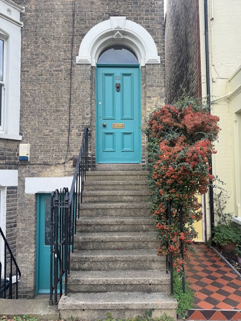The image depicts the exterior of a residential building with a set of concrete stairs leading up to a bright blue front door, which is framed by a white archway. To the right of the stairs, there is a tall shrub with reddish-green leaves, partially covering a black metal railing that runs alongside the steps. The building has a brick facade, and a small basement window with a blue frame is visible at the bottom of the staircase. The surrounding area shows a paved pathway with a red and black checkered pattern adjacent to the building. The lighting suggests daytime, with natural light illuminating the scene. This setting illustrates typical elements encountered in house relocations involving narrow doorways and stairs, a common challenge for removals and furniture transport companies like Removal Company Hounslow, especially during home relocation or furniture moving procedures.