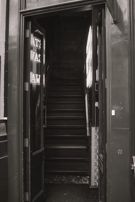 This image depicts a narrow, steep staircase inside a building, constructed from worn stone or concrete with visible cracks and uneven surfaces. Metal handrails run along both sides of the staircase, attached to the walls which are painted in a faded light color with signs of age and dirt. At the top of the stairs, there is a small landing area illuminated by a single exposed yellowish light bulb, casting warm light on the wooden door and the surrounding wall. The door appears to be slightly ajar, providing a glimpse into another room beyond. The overall scene reflects the challenges of home relocation involving tight spaces and narrow doorways, which are often encountered during furniture transport or packing and moving processes, where careful navigation of confined areas is necessary. Removal Company Hounslow staff would carefully manage such spaces during internal or external moving tasks to ensure safe and efficient relocation of household items.
