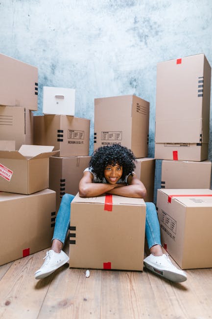 A young woman with curly black hair and a bright smile is sitting on the wooden floor inside a room, surrounded by numerous packed cardboard boxes of various sizes, some sealed with red or black tape. She rests her chin on a large box in front of her, with her arms draped over it, and is dressed in a white top, blue jeans, and white sneakers. The walls of the room are painted in a light, textured blue, and the space appears to be prepared for a home relocation or moving process. The boxes are stacked against the walls, indicating an organized packing and moving setup, with some boxes partially open or labeled. This scene captures the packing and moving stages handled by a professional removals service, such as Removal Company Hounslow, in preparation for a house relocation.