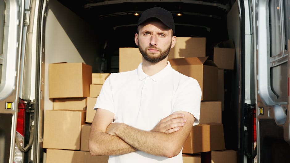 A young man with a serious expression, wearing a white polo shirt and black cap, standing inside the back of a moving van surrounded by stacked cardboard boxes and packing materials. The boxes vary in size, some wrapped in plastic or packing tape, and are positioned on the floor and shelves of the van. The interior of the van is dark, with the open rear doors allowing natural light to illuminate the scene. In the background, additional boxes are visible, along with loading equipment such as pallets or trolleys. This scene reflects the typical home relocation process involving furniture transport, packing, and loading for a house removal service, as provided by Removal Company Hounslow, supporting smooth logistics and moving operations.