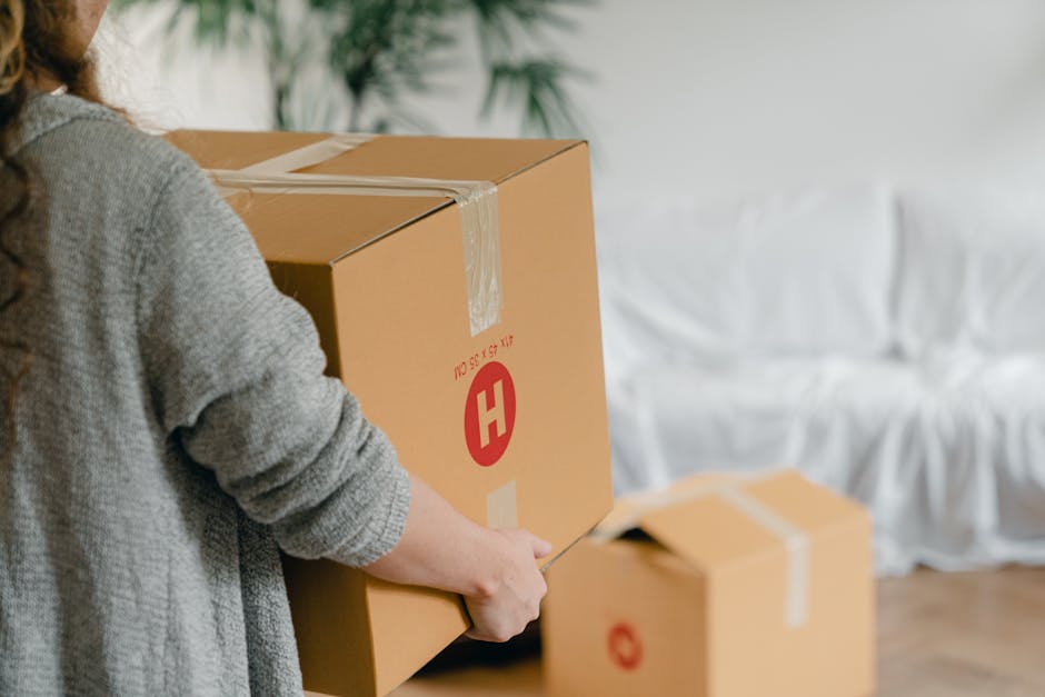 A person wearing a grey sweater is in the process of packing a large cardboard moving box, which features a red 'H' logo and printed measurements, in an indoor setting with white walls and a sofa covered with a white slipcover in the background. The individual is using a utility knife to seal the top of the box, positioned on a wooden floor near other packed boxes. Several boxes are visible, some already sealed with packing tape, indicating a home relocation or furniture transport process. The scene captures the detailed packing stage of a house removal, with cardboard boxes and packing materials such as tape being used to prepare belongings for moving services. The lighting is natural, highlighting the organized setup for a scheduled move, associated with house removals services provided by Removal Company Hounslow.